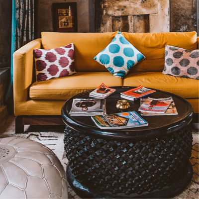 Yellow couch with 3 poka-dotted pillows on it and a giant round coffee table covered in magazines.
